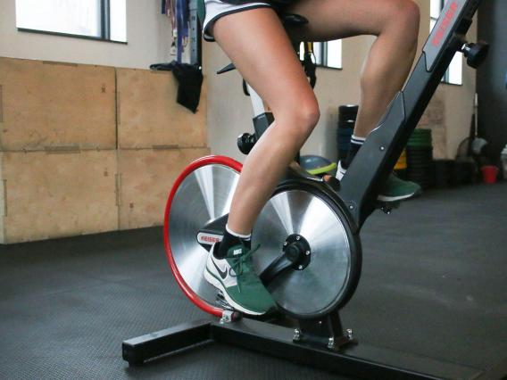 A waist-down shot of a cyclist in shorts and athletic shoes pedaling a stationary bicycle in an indoor setting.